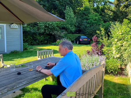 Before image shows a silver people carrier car to the left on a gravel drive in front of a garage. Large wooden table and chairs are to the right on a large grassed area. Low hedges are featured around the boundary of the garden.There are lime green sketch marks on the images showcases ideas for improvements of flower beds and bushes. the after image shows a man sitting on a wooden bench at a wooden table over looking the garage. There are planted borders to the right featuring greens, dark and light purple flowers. A black sports car is now parked on the grass to the far side fo the garden, behind the garage. Click image for more details.