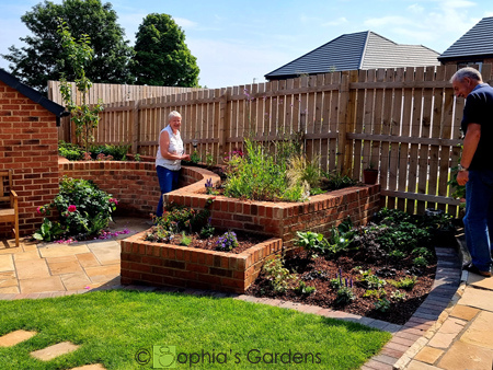 Before image shows a red brick garage wall with natural wood fence surrounding the rear of the garden. The ground is just a layer of mud, with no grass or planting. a blue bin is visible at the forefront of the image. the after image shows a lady and a man looking over lovely planting in  raised, curved beds now situated in front of the natural wood fence. The brick walls for the beds are the same red as the garage wall. The ground now features a beautiful natural stone patio to the left , in front of the garage, and grassed areas to the rest. With stepping stones over the grass. Click image for more details.