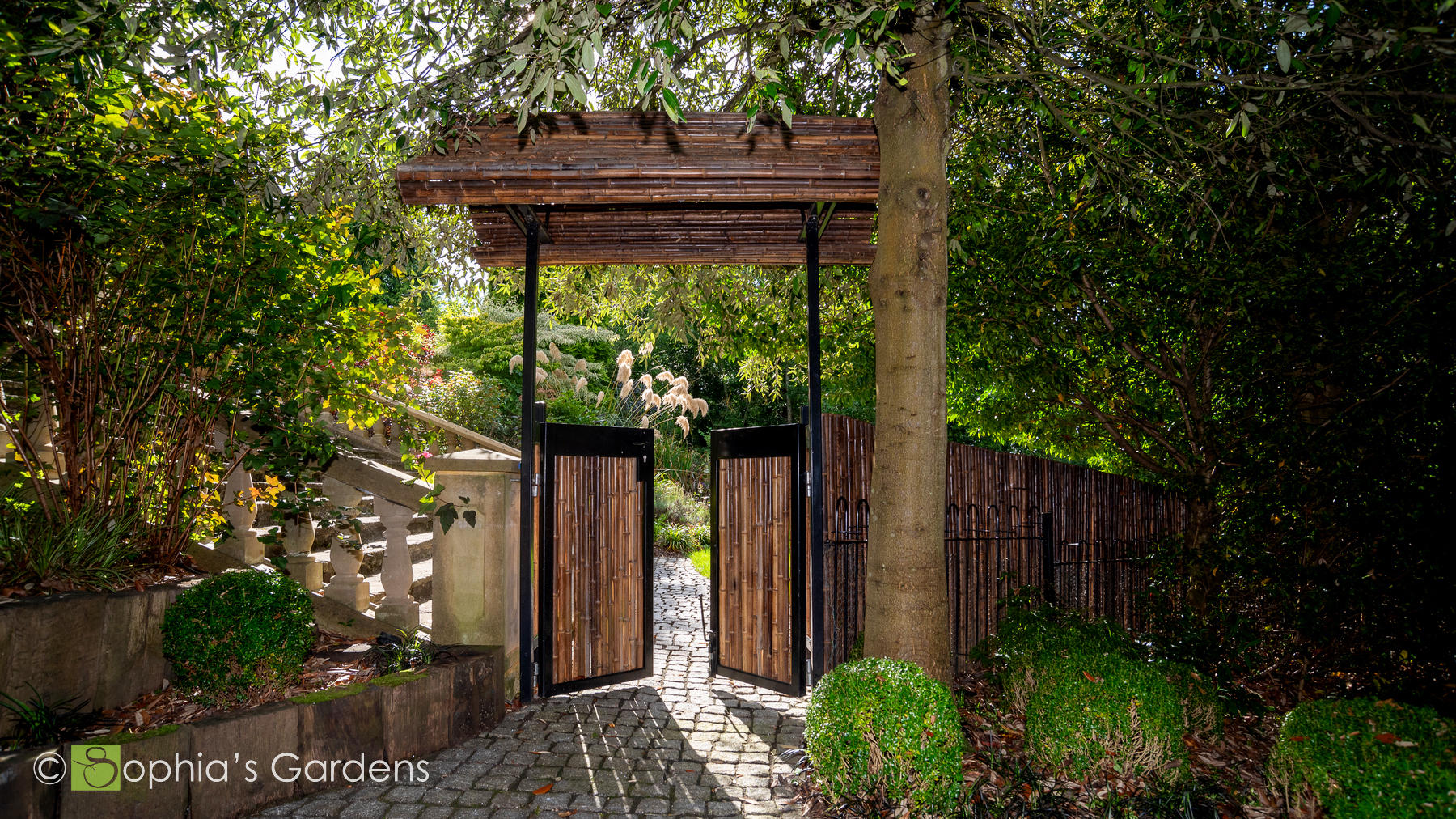 Japanese gate made with bamboo and cast iron with a bamboo arch, looking through to grasses in an ornamental flowerbed, in Darras Hall, Ponteland, by Sophia's Gardens