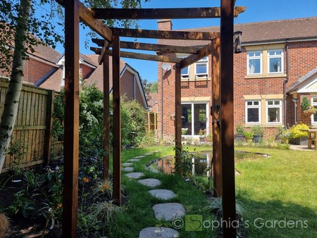 Image of a curved pergola over a curvy stepping-stone path leading past a wildlife pond to a patio in front of a modern-traditional house