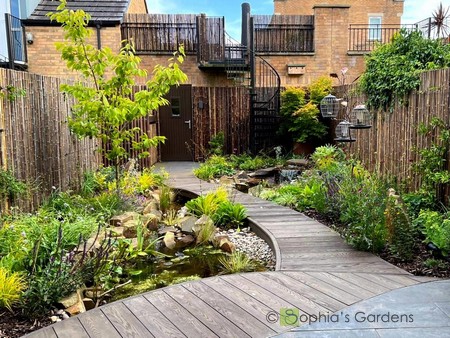 A basic rectangular garden transformed into a lush Japanese-inspired watery wonderland. A decking path leads diagonally across the garden and over a stream which runs between a waterfall and a wildlife pond.